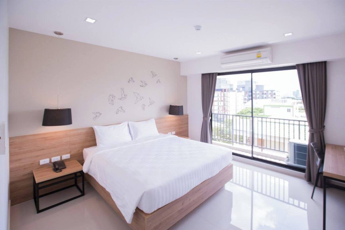 Una habitación de hotel moderna con una cama grande, una mesita de noche con lámpara, aire acondicionado en la pared y vista a la ciudad desde un balcón.