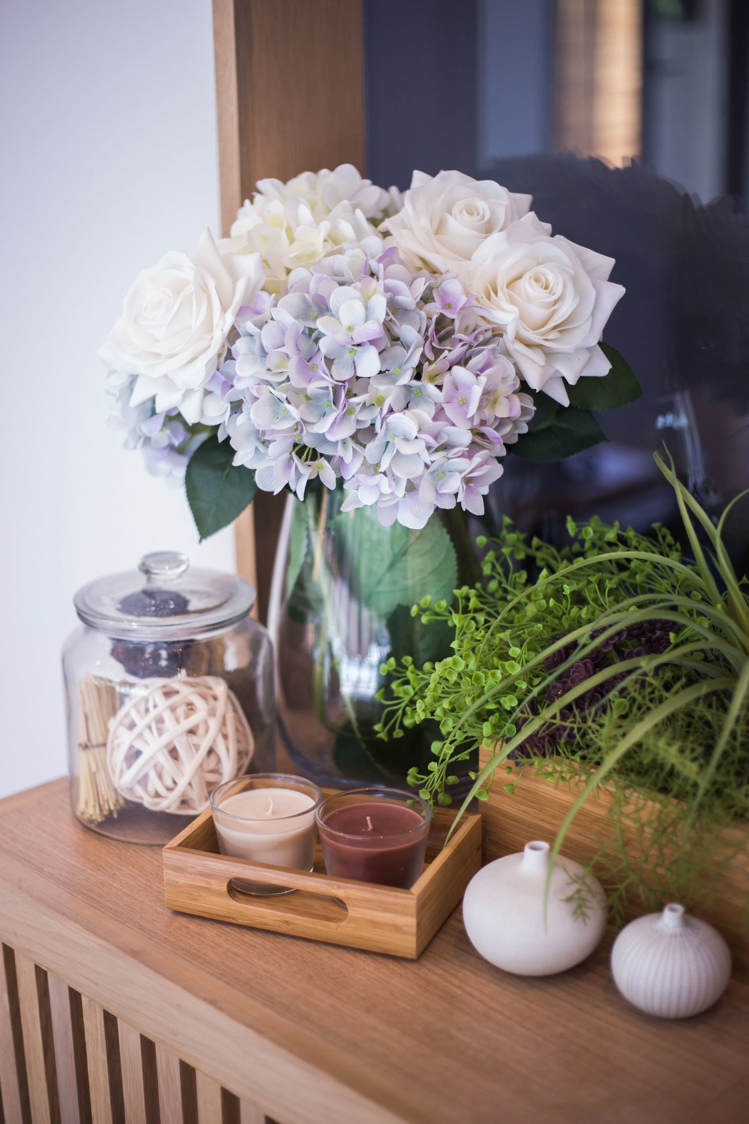 Una mesa de madera con un jarrón de rosas blancas e hortensias, un tarro de cristal y pequeños jarrones.