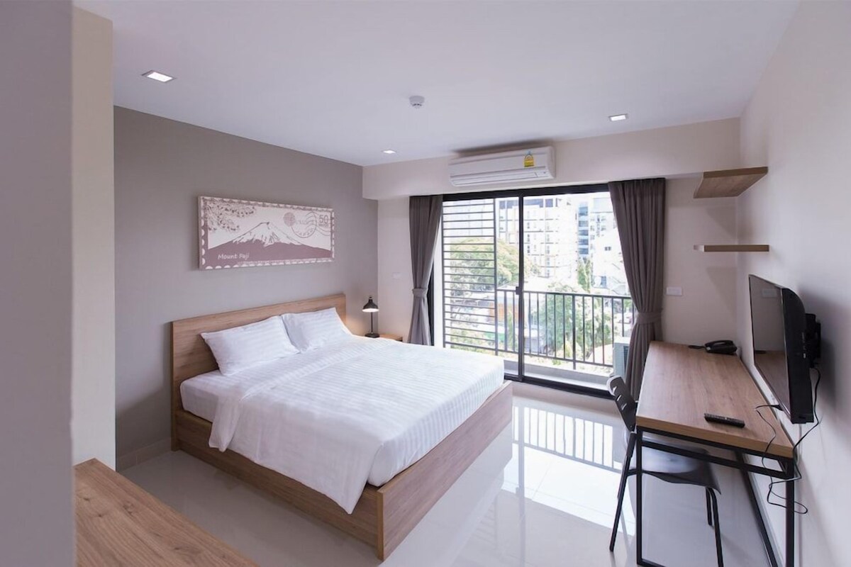 A modern hotel room with a bed, a desk with a chair, a television, and a balcony with a view of buildings.