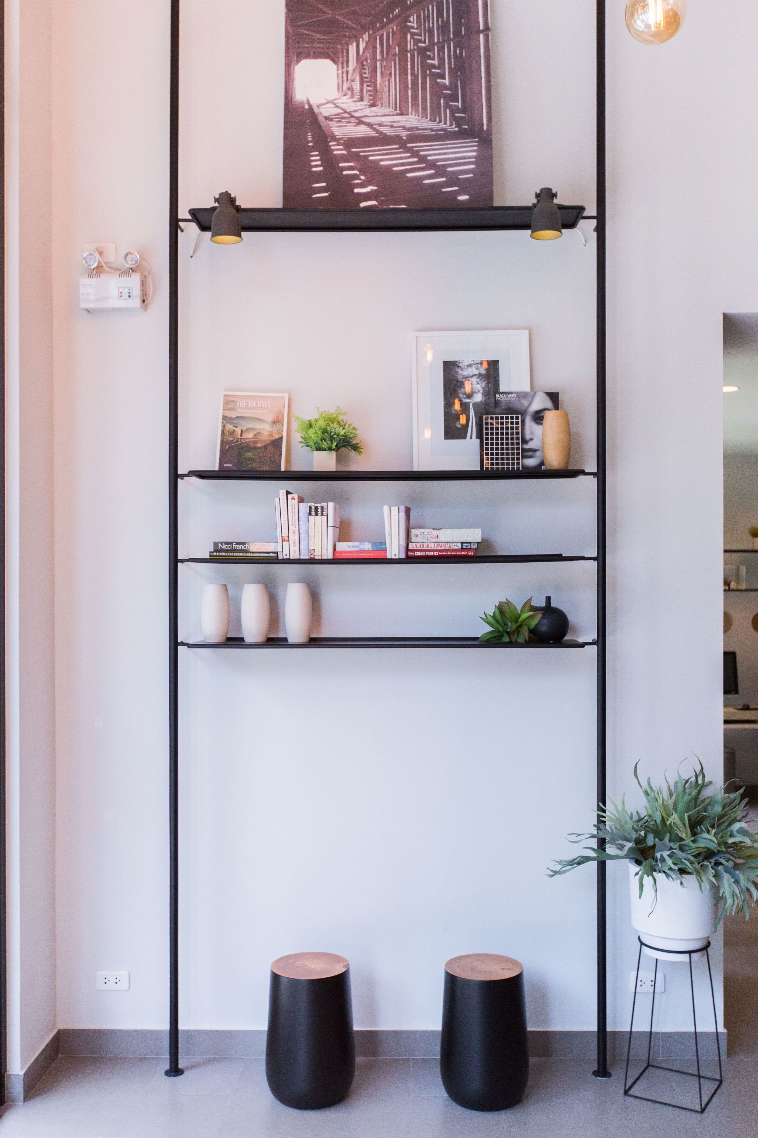Un intérieur moderne avec une étagère en métal noir où sont exposés des livres, des objets décoratifs et un tableau encadré. On y trouve également deux tabourets noirs et une plante en pot sur le sol.