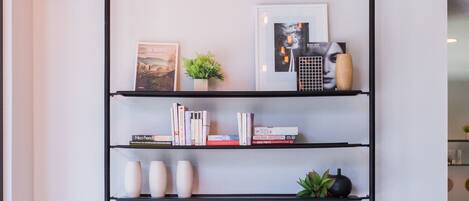 A modern interior with a black metal shelving unit displaying books, decorative items, and a framed picture. There are two black stools and a potted plant on the floor.