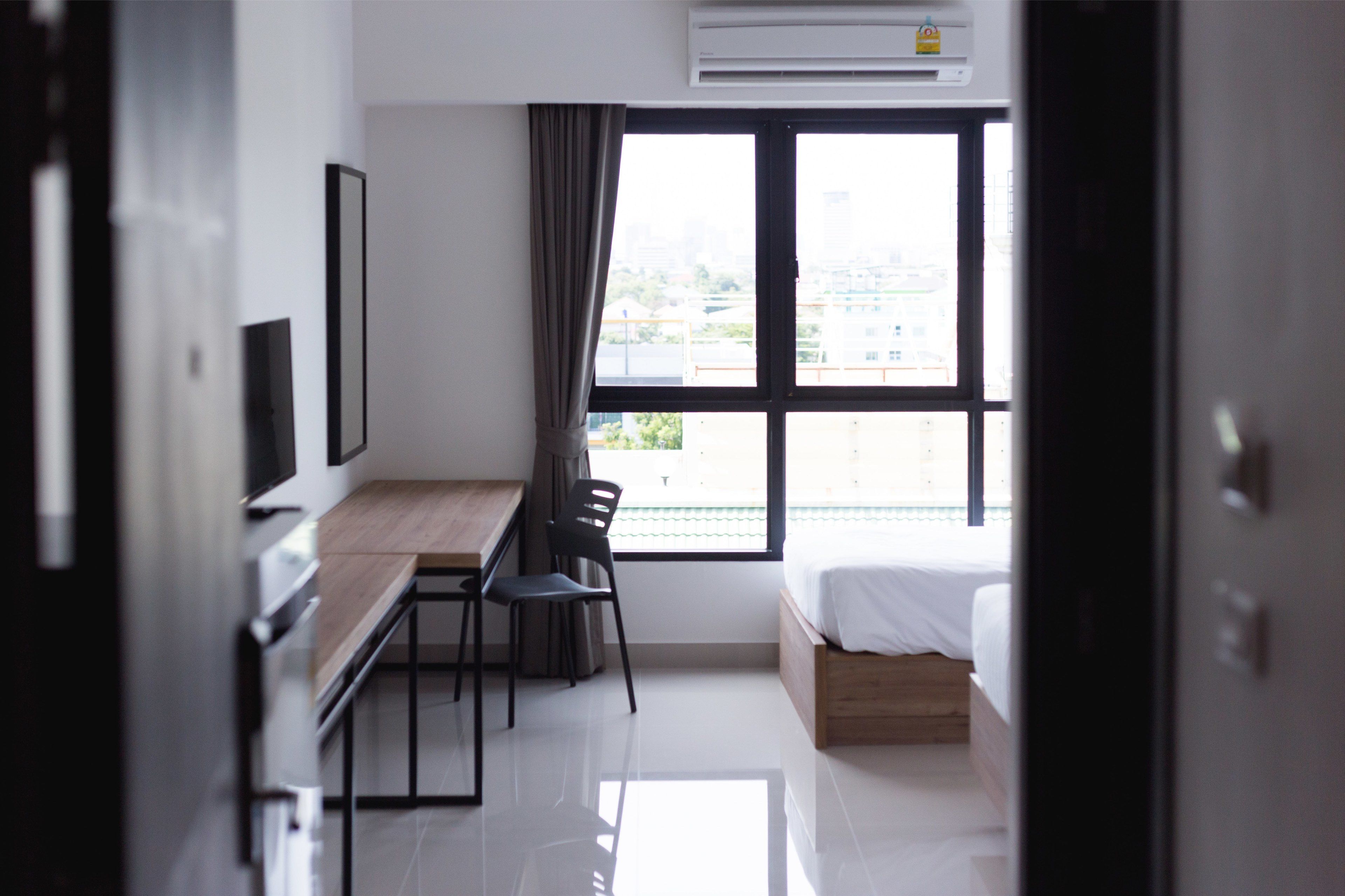 A modern hotel room with a wooden desk, a chair, a bed, and a large window with a city view.