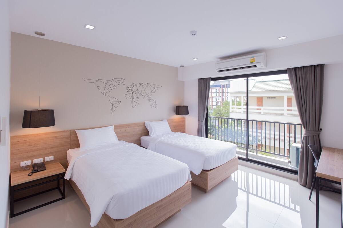 Habitación de hotel con dos camas, un escritorio, un mapa mundial en la pared y un balcón con vistas.