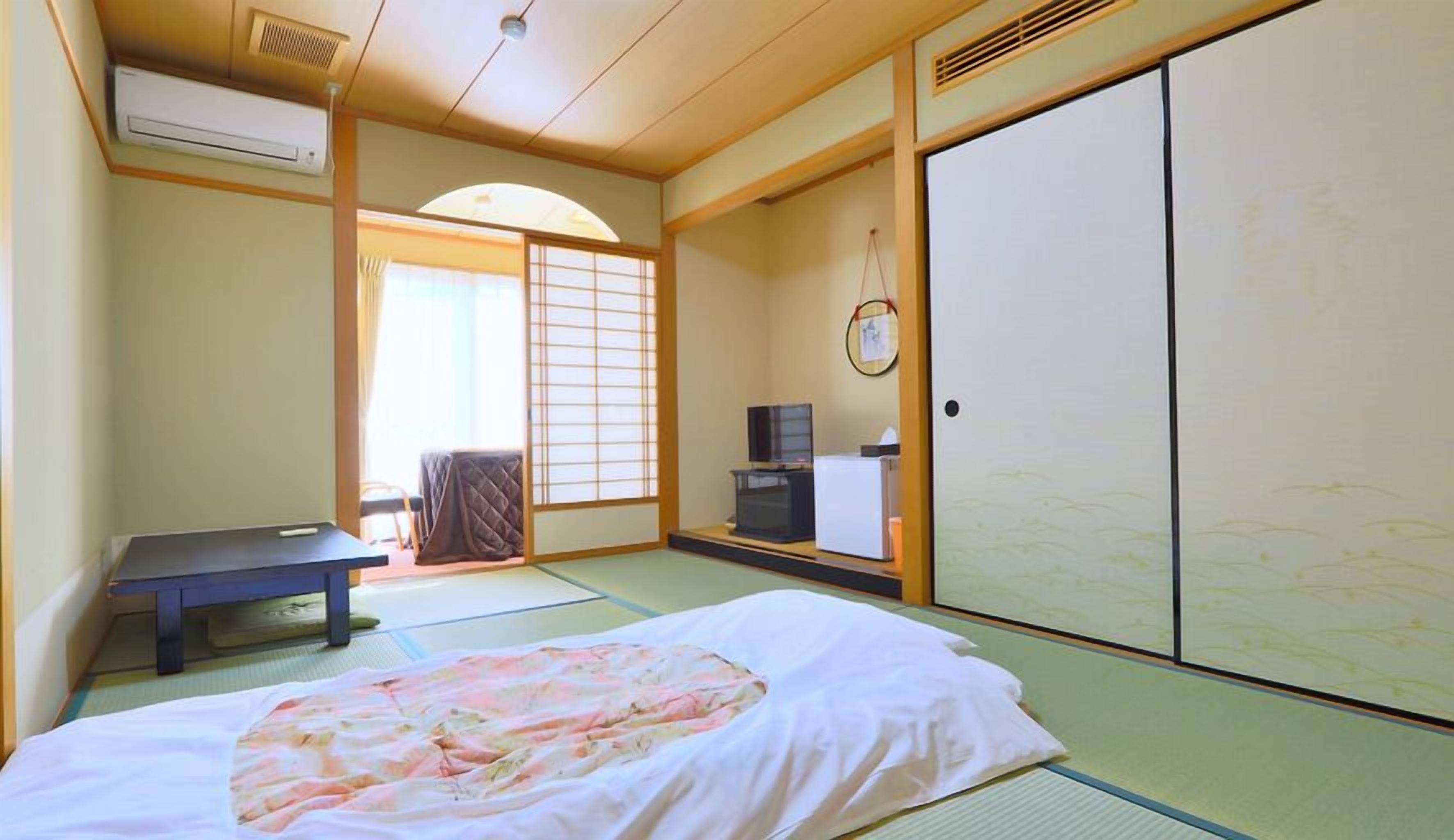 Une pièce de style japonais traditionnel, avec un sol recouvert de tatamis, un lit avec une couvre-lit à motifs floraux, un bureau, une télévision et des portes coulissantes.