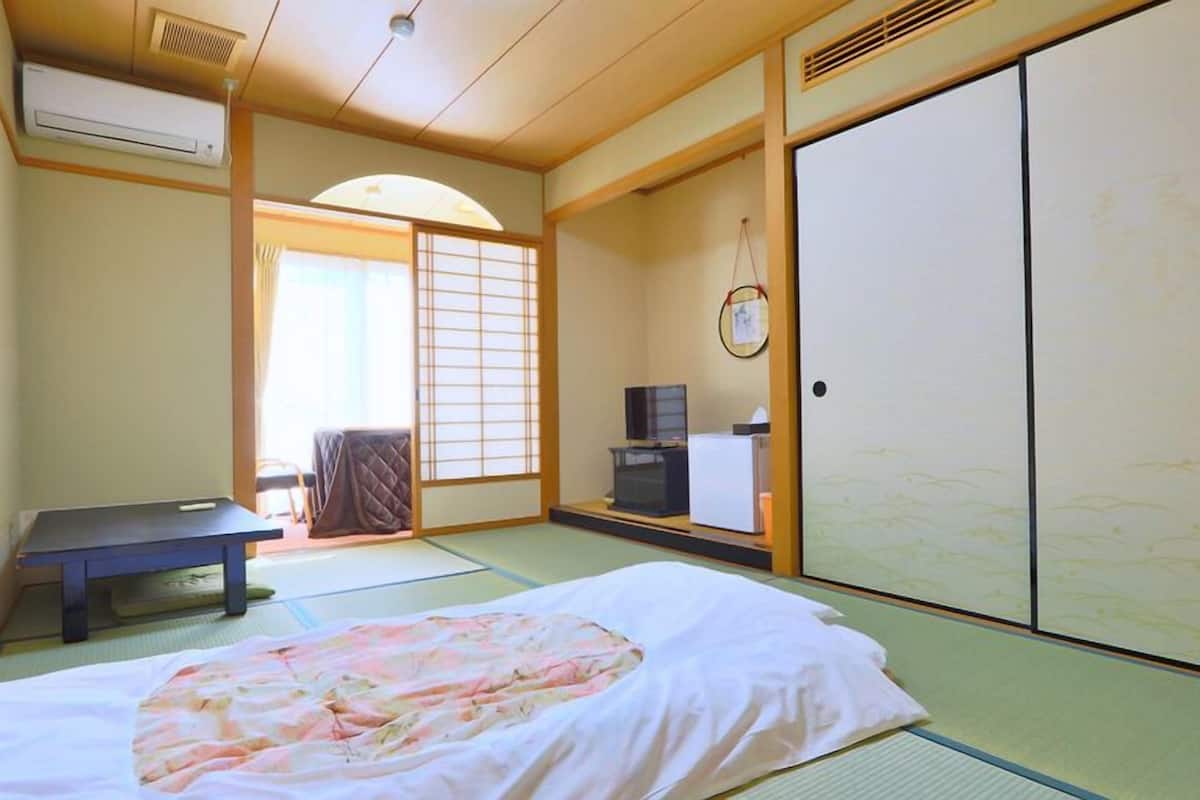 Une pièce de style japonais traditionnel, avec un sol recouvert de tatamis, un lit avec une couvre-lit à motifs floraux, un bureau, une télévision et des portes coulissantes.