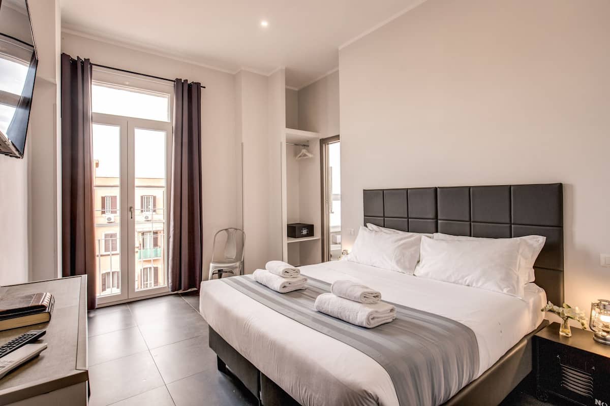 A hotel room with a large bed, a desk with a chair, a TV mounted on the wall, and a window with curtains.