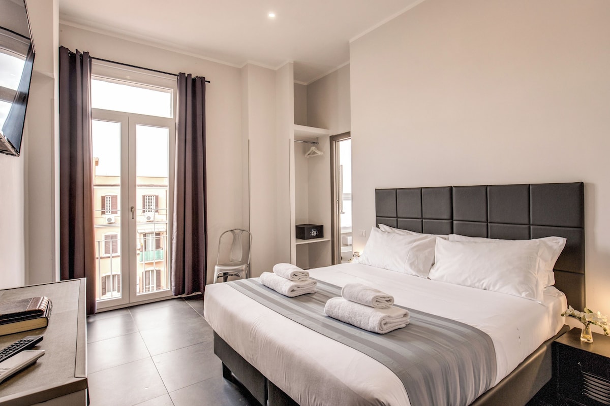 A hotel room with a bed, a desk, a chair, a TV, and a view of the outside.