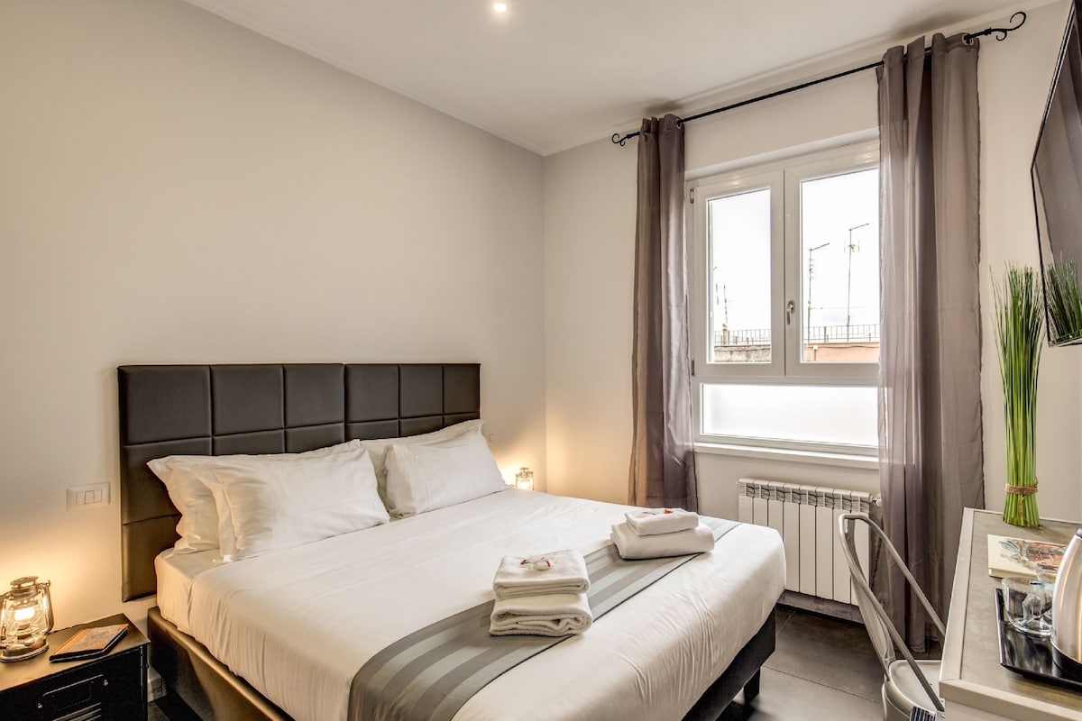 A neatly made bed with white linens and a gray striped bedspread, a bedside table with a lamp, and a window with curtains.