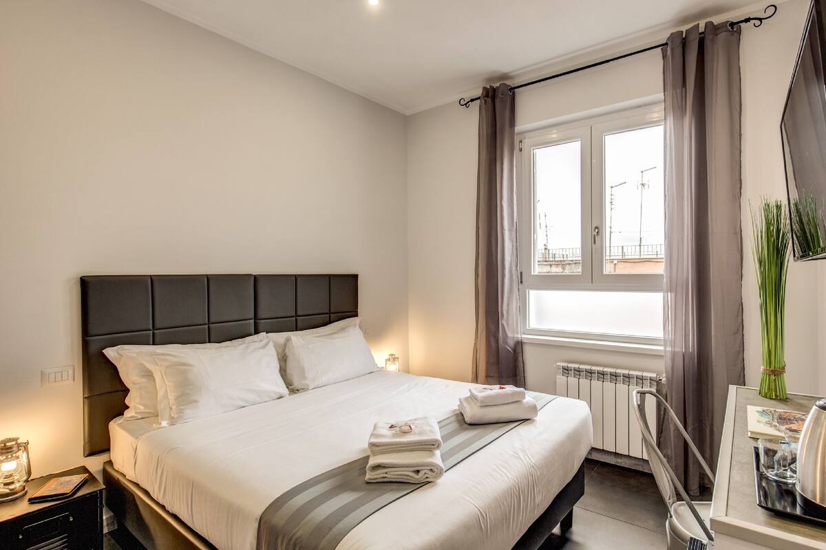 A neatly made bed with white linens, a bedside table with a lamp, and a window with curtains.