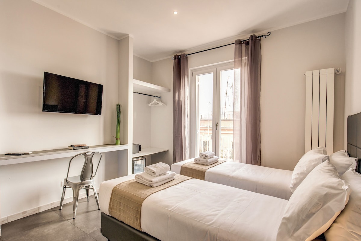 A hotel room with two beds, a desk, a chair, a TV, and a window with curtains.