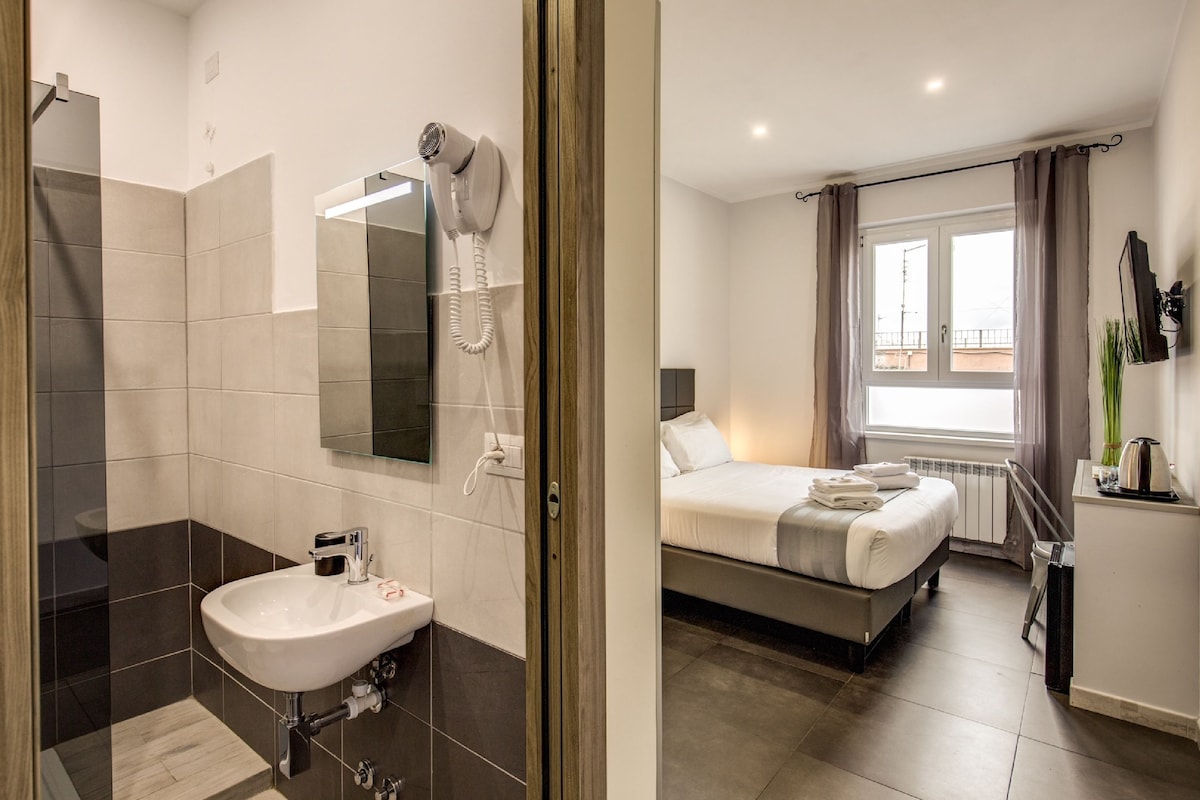 A modern bathroom with a shower, sink, and hairdryer. The room has a bed with a view of the outdoors through a window.