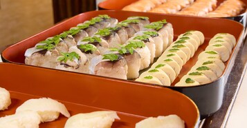 Un bufé con distintos alimentos, como pescado en rodajas y panecillos, dispuestos en bandejas.