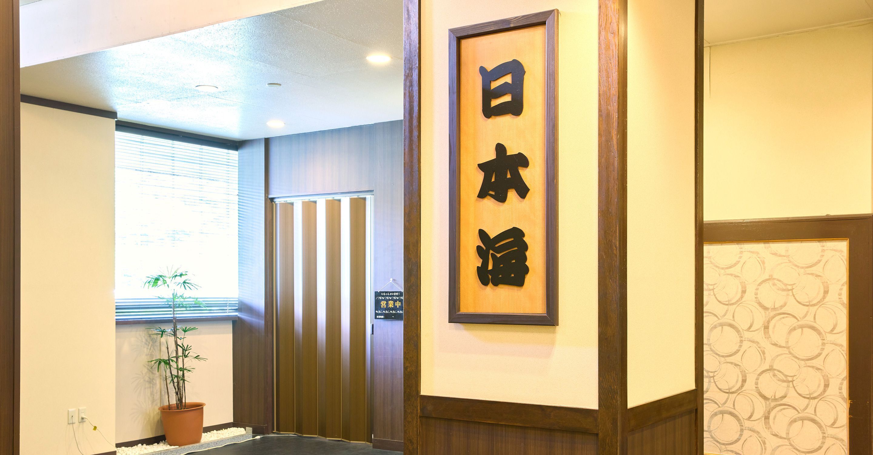 A reception area with a wooden sign with Japanese characters, a potted plant, and a door with vertical blinds.