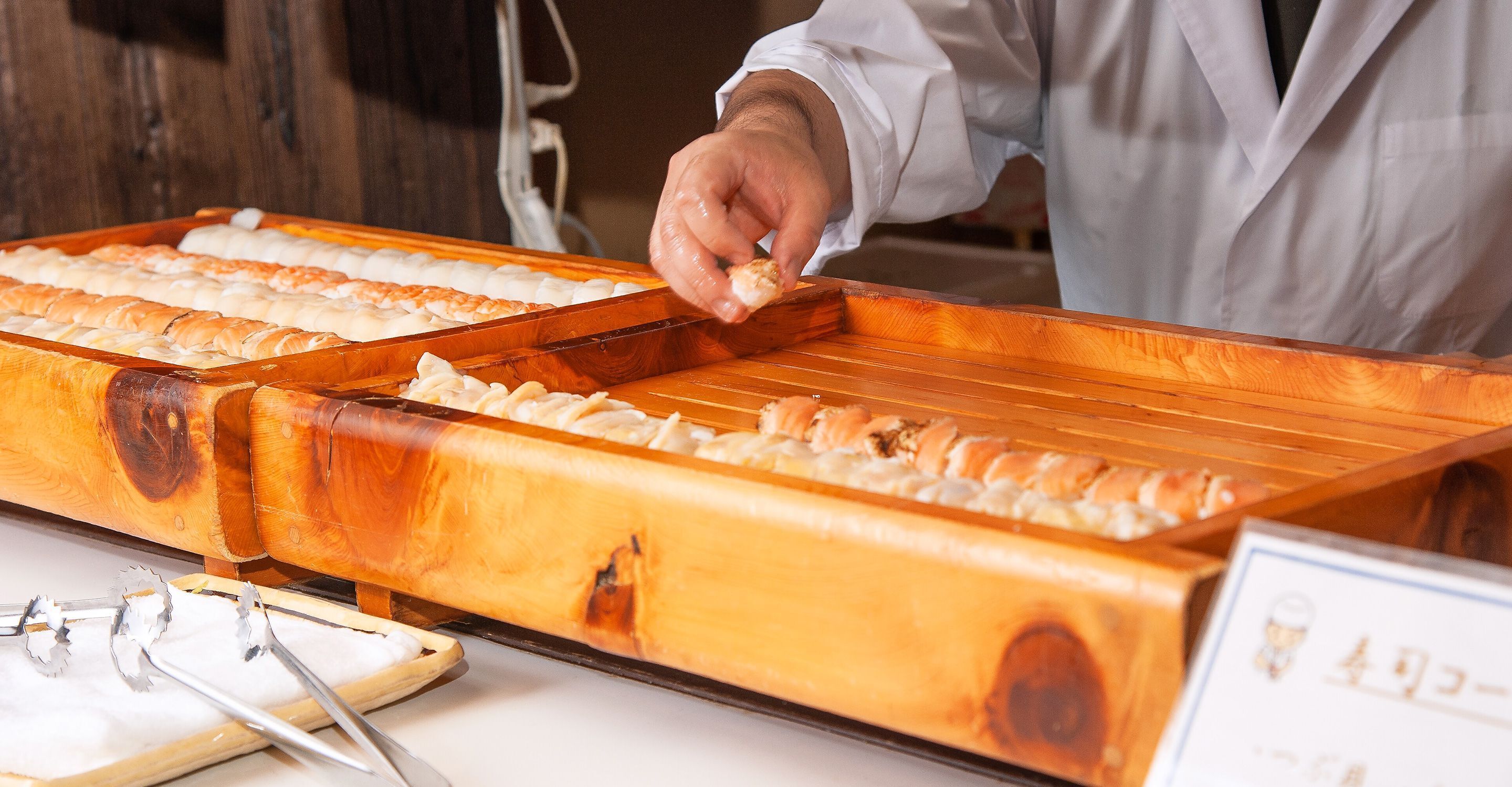 A person preparing sushi at a wooden counter with multiple trays of sushi.