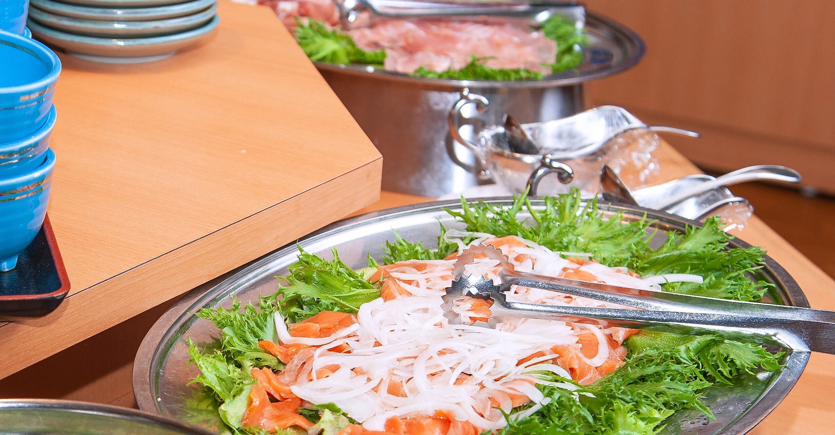 Una mesa de comedor con un bol metálico que contiene salmón en rodajas y verduras ralladas, un par de pinzas y una pila de boles azules.