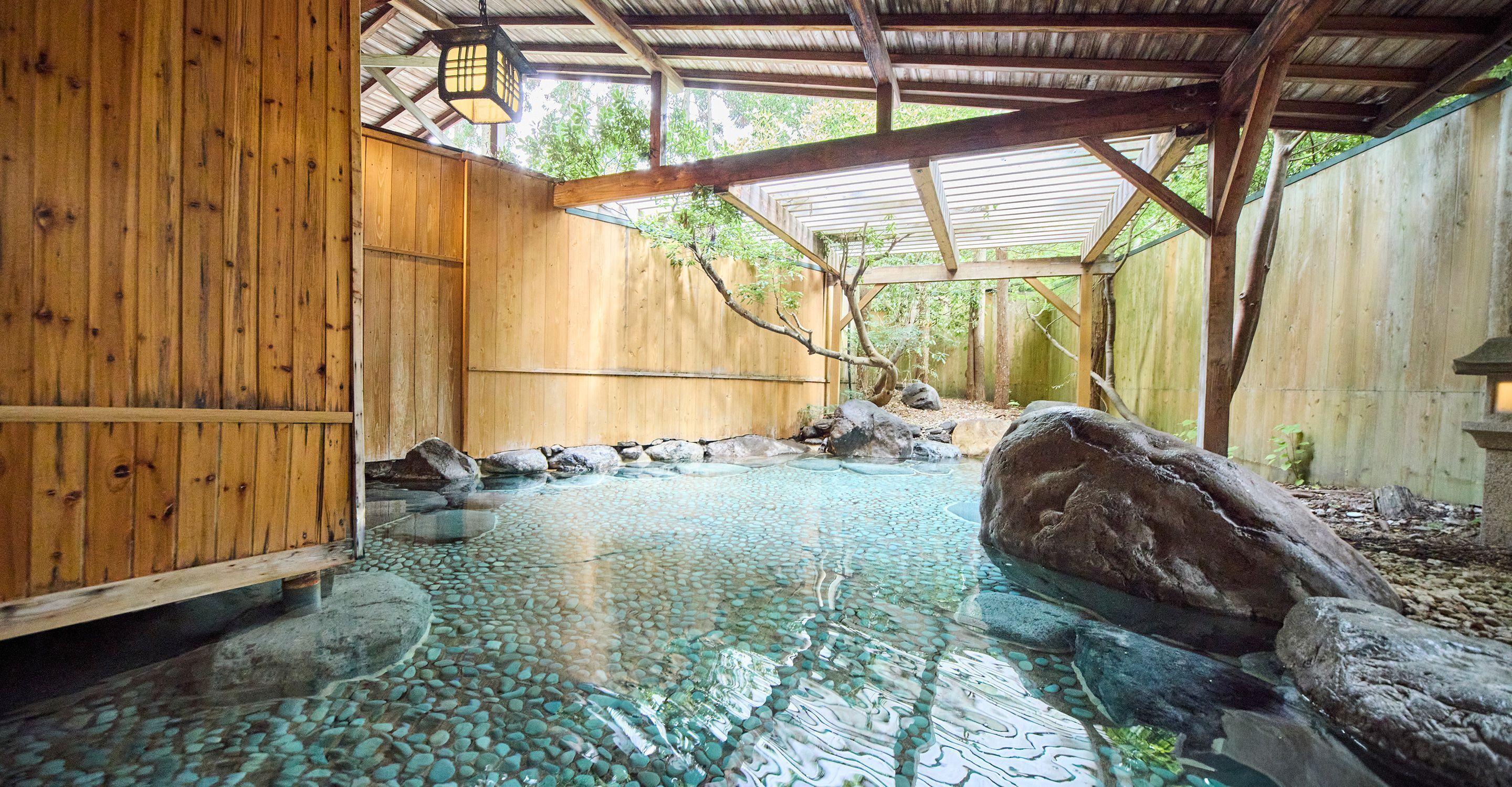 A traditional wooden bathhouse with a clear stone pool, wooden benches, and a rustic interior.