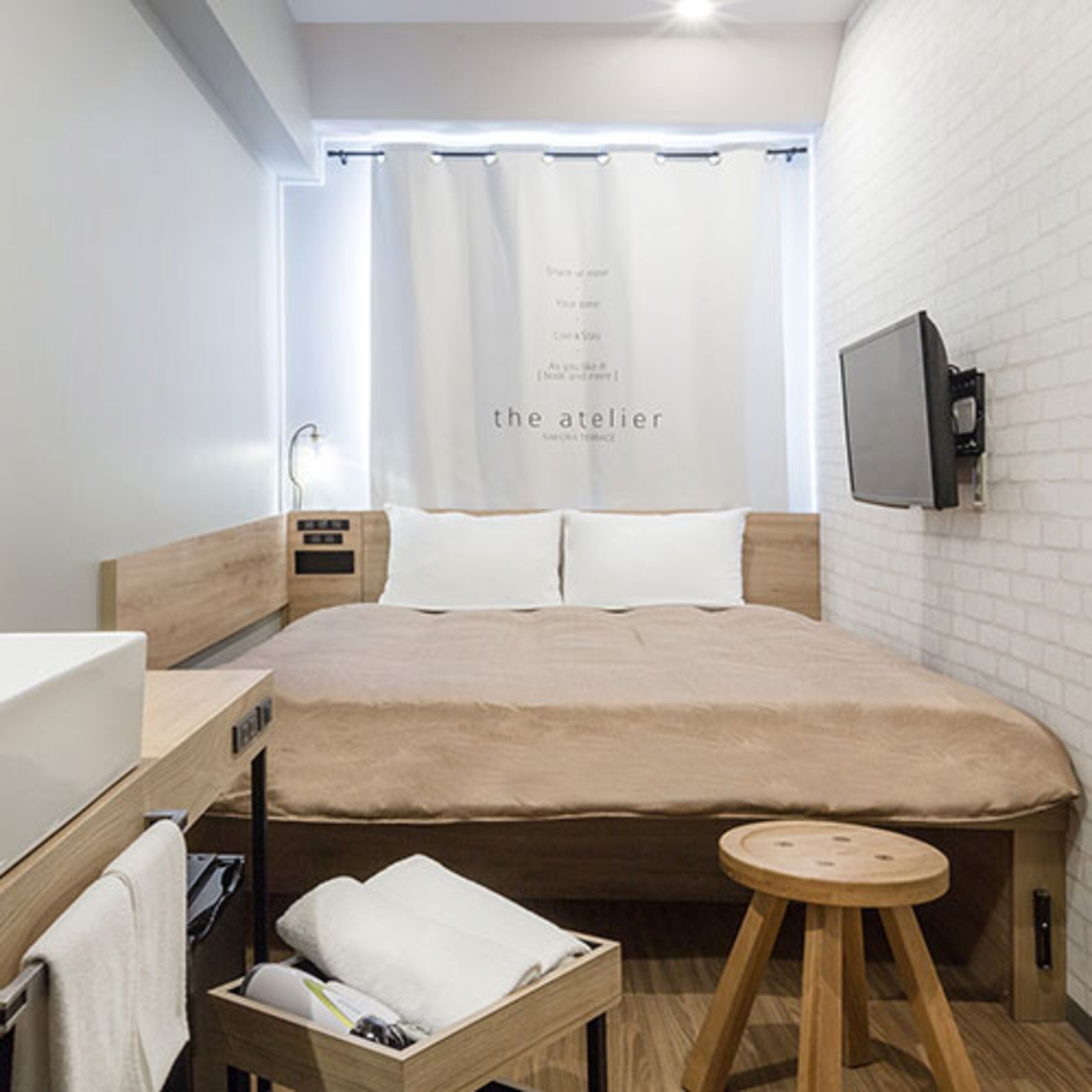 Un dormitorio moderno con una cama grande, una mesita de noche, un taburete pequeño, un televisor montado en la pared y una ventana con cortina.