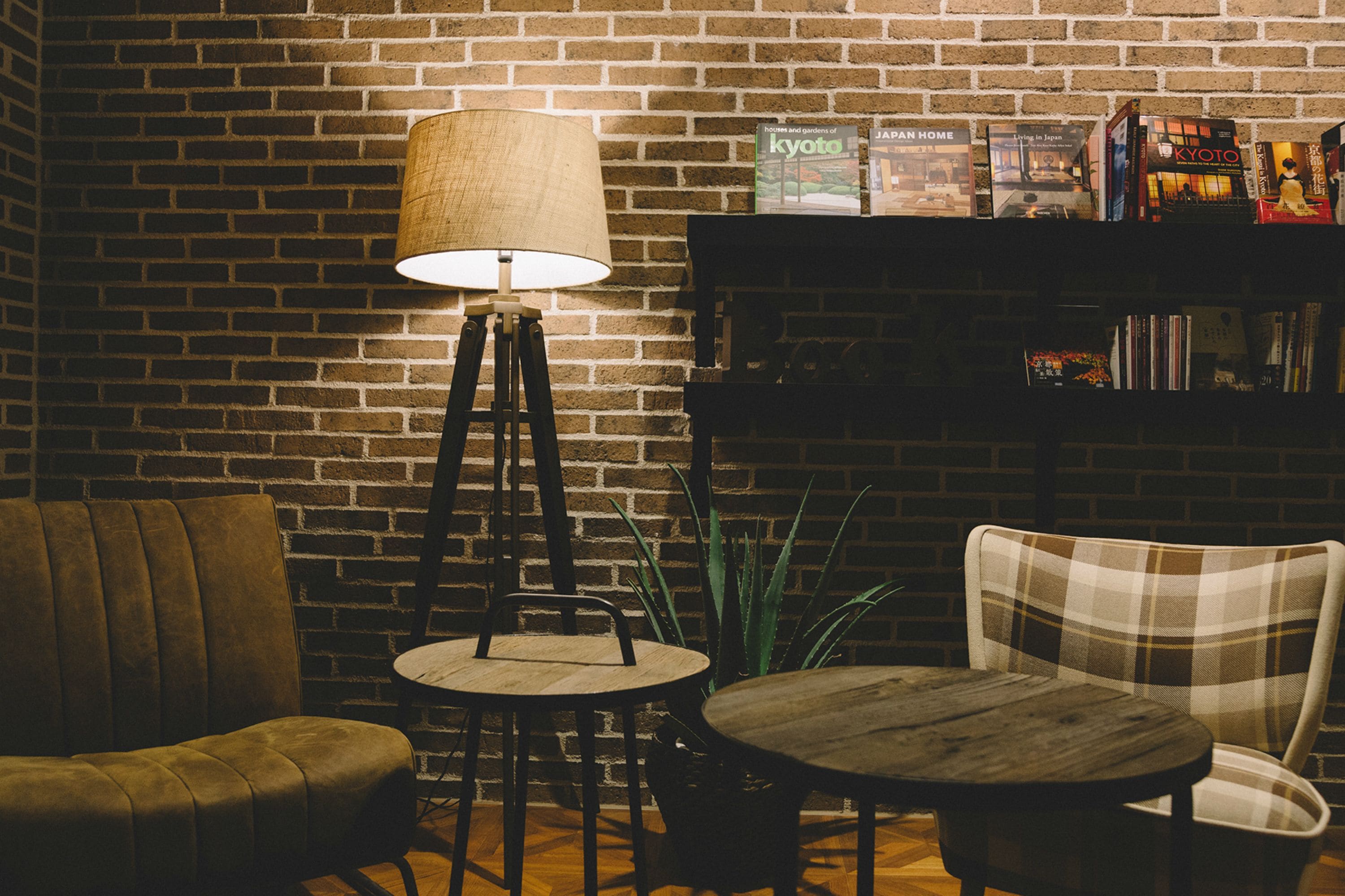 A cozy seating area with a brick wall, a floor lamp, a bookshelf with books, and a potted plant.