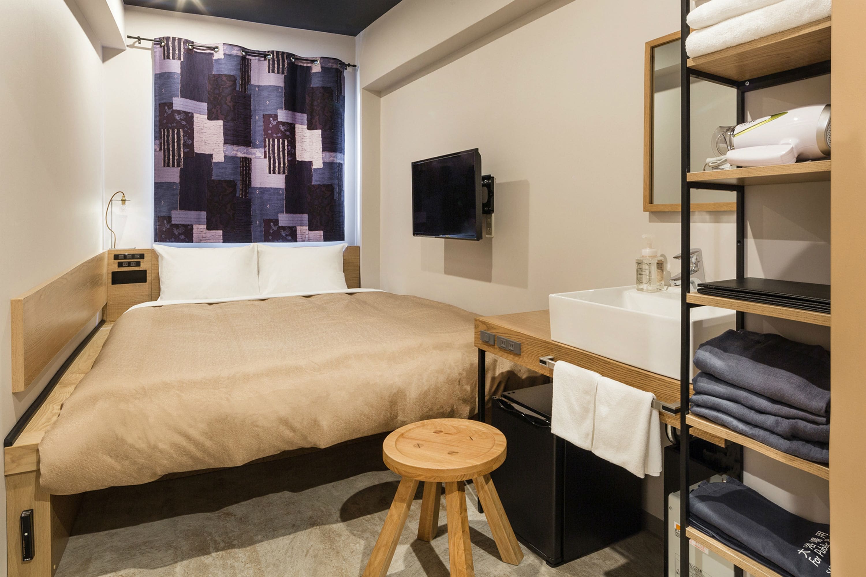 A hotel room with a bed, a wooden stool, a desk with a sink, a mirror, and shelves with towels and folded clothes.