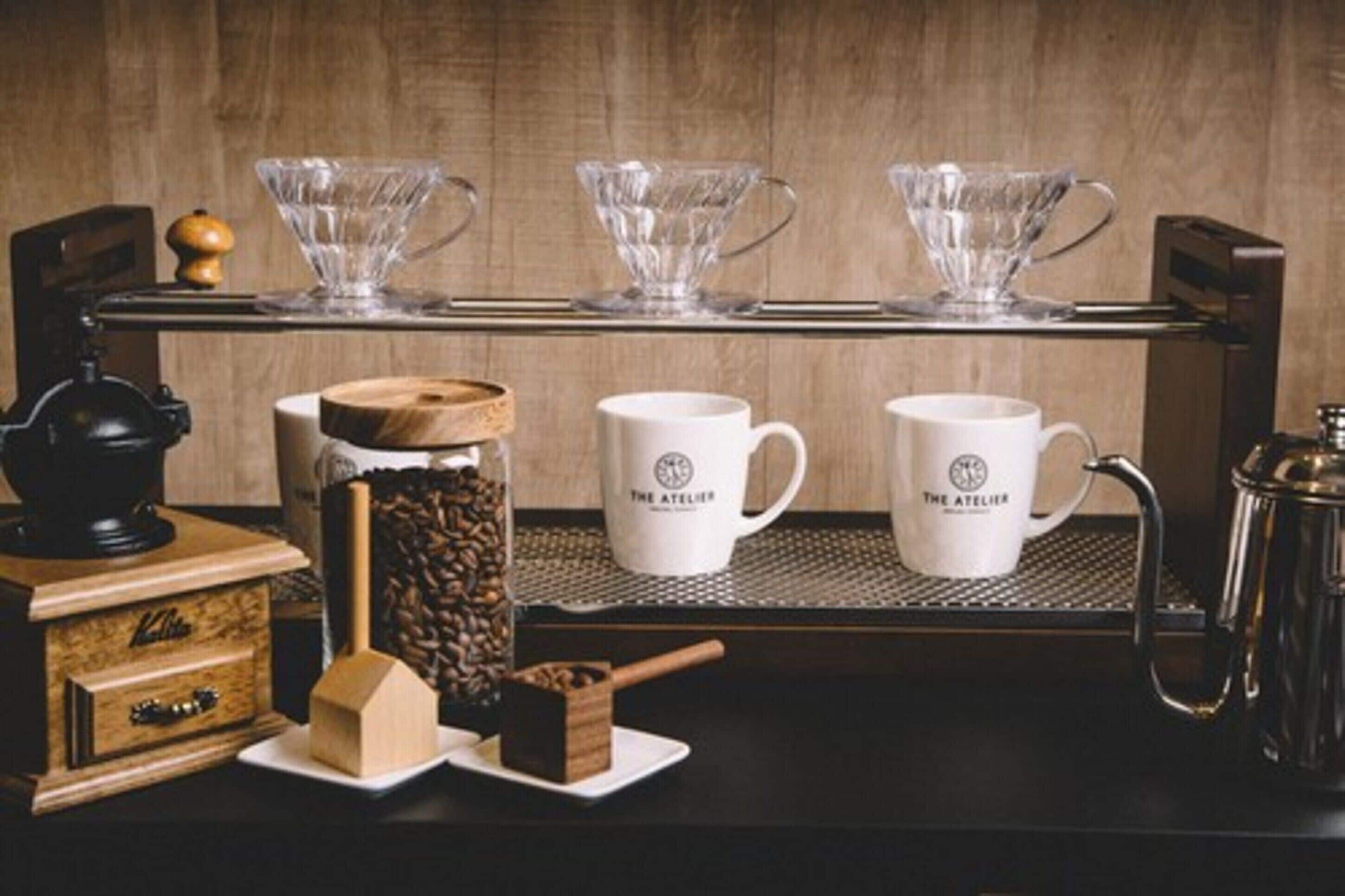 Eine Kaffeestation mit einer Kaffeemühle, einem Pressstempel und zwei weißen Tassen mit dem Aufdruck 'The Atelier'.