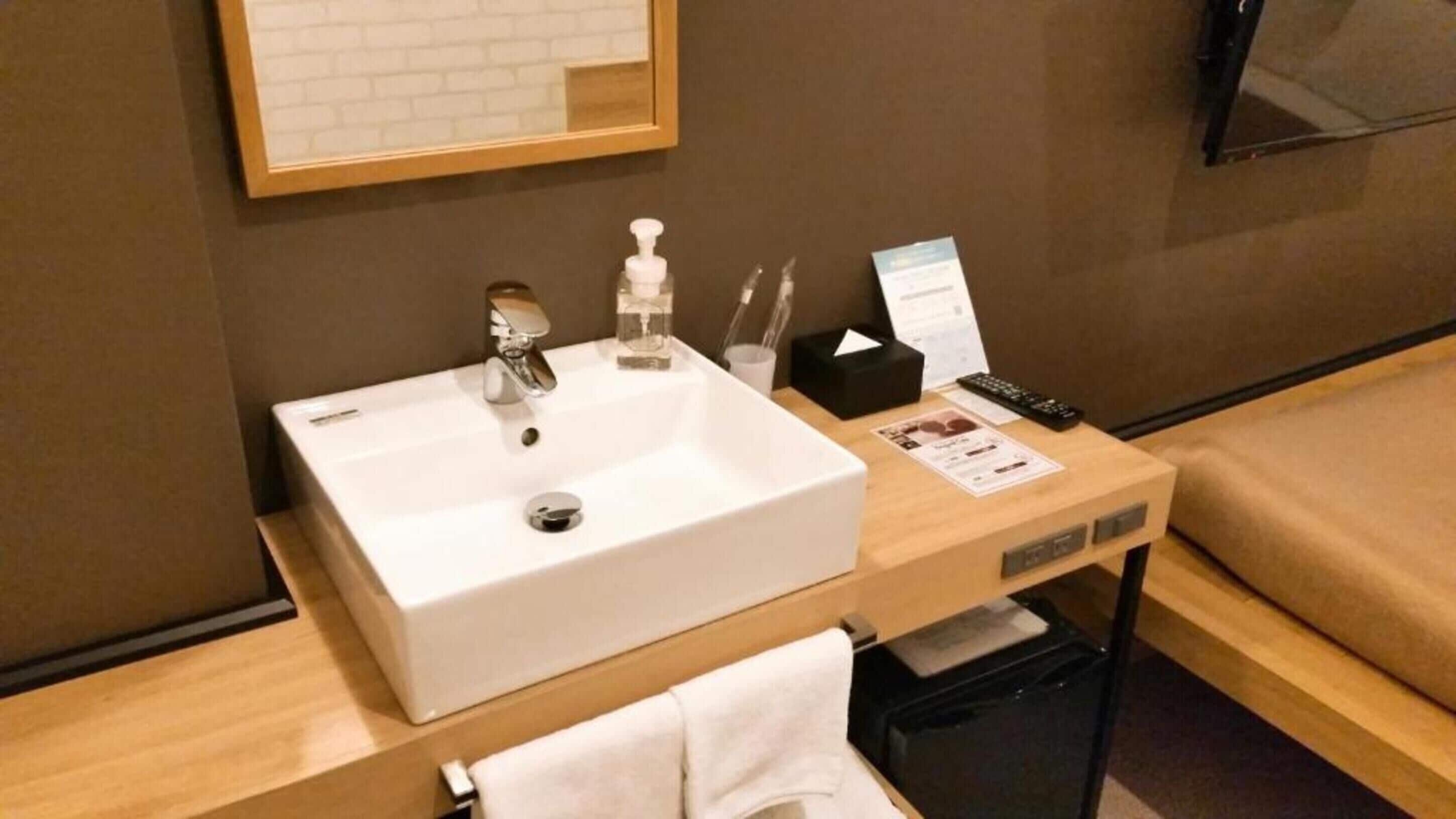 A hotel bathroom with a white sink, a mirror, a bottle of hand soap, a tissue box, and a remote control on a wooden side table.