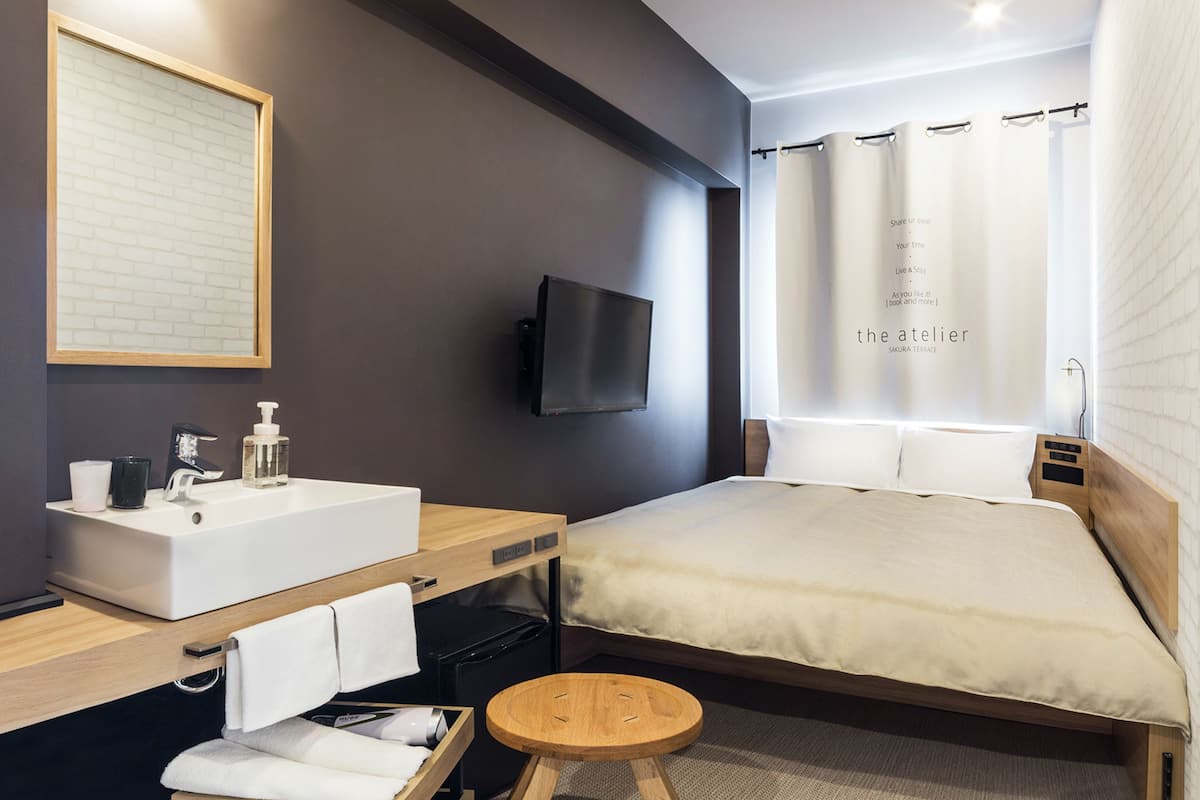 A modern hotel room with a large bed, a bedside table with a lamp, a TV mounted on the wall, and a window with curtains.