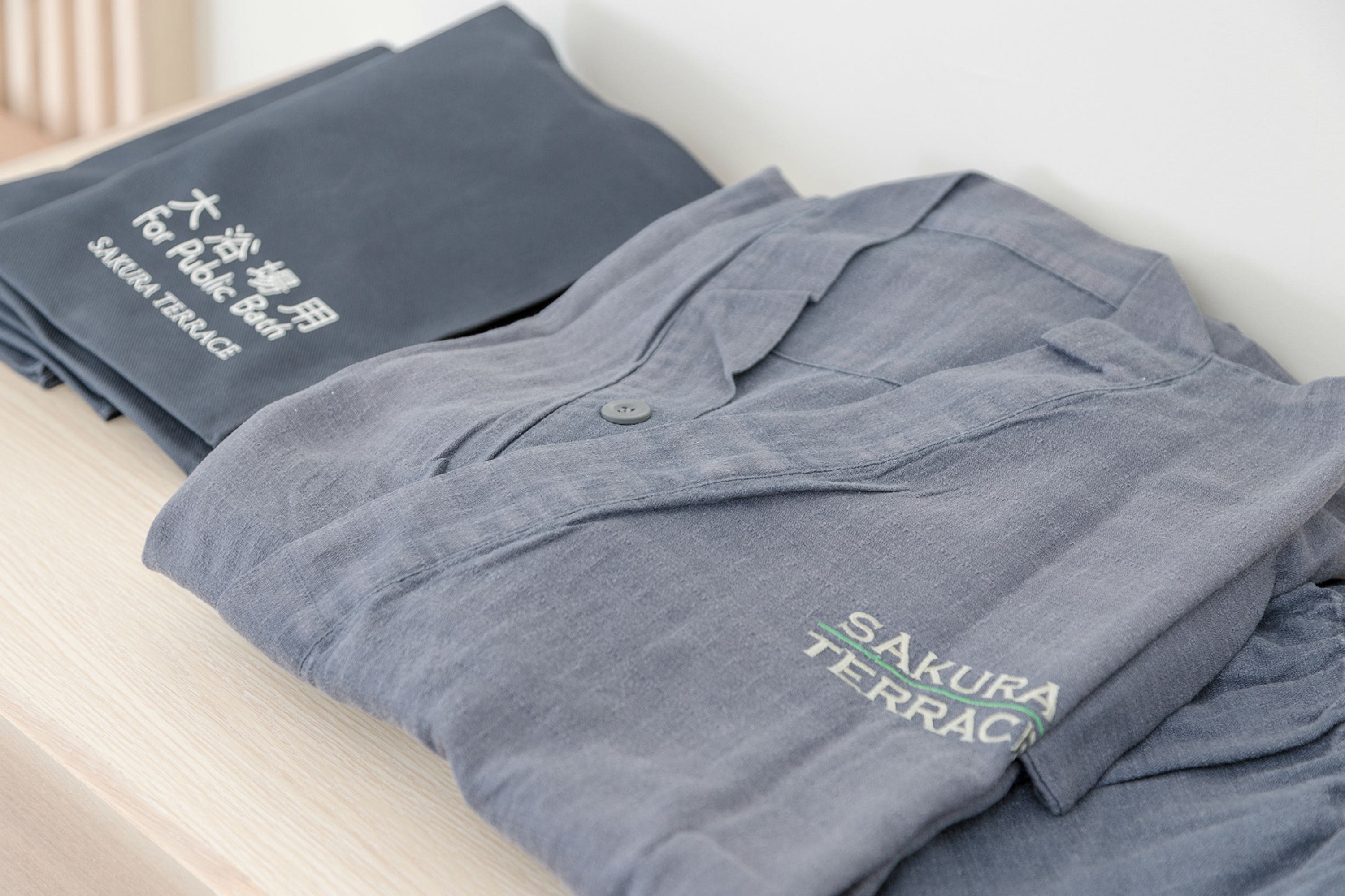 A neatly folded grey shirt with "SAKURA TERRACE" embroidered on it, placed on a wooden surface.