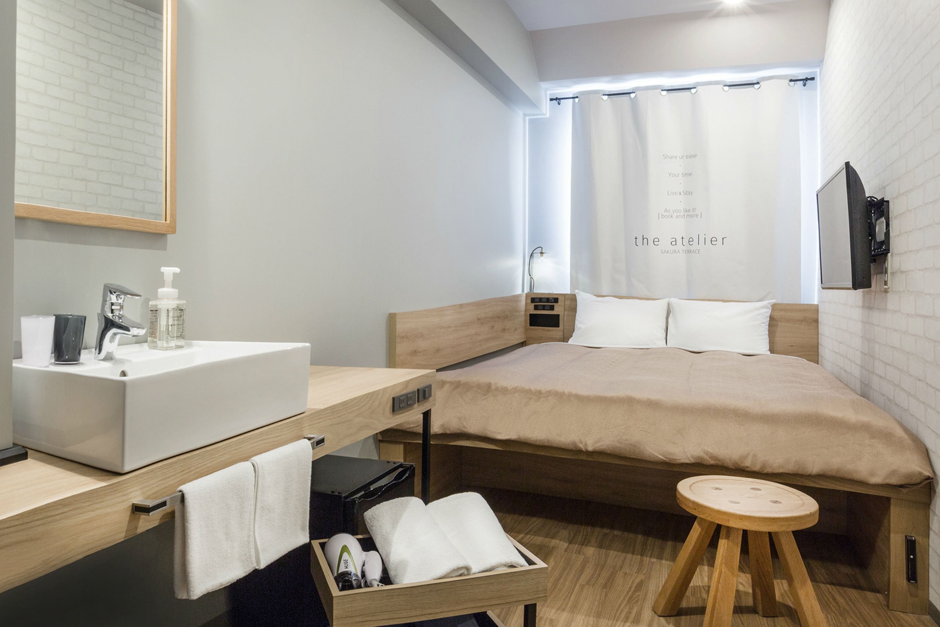 A modern hotel room with a wooden bed, a built-in desk with a sink, and a small stool.