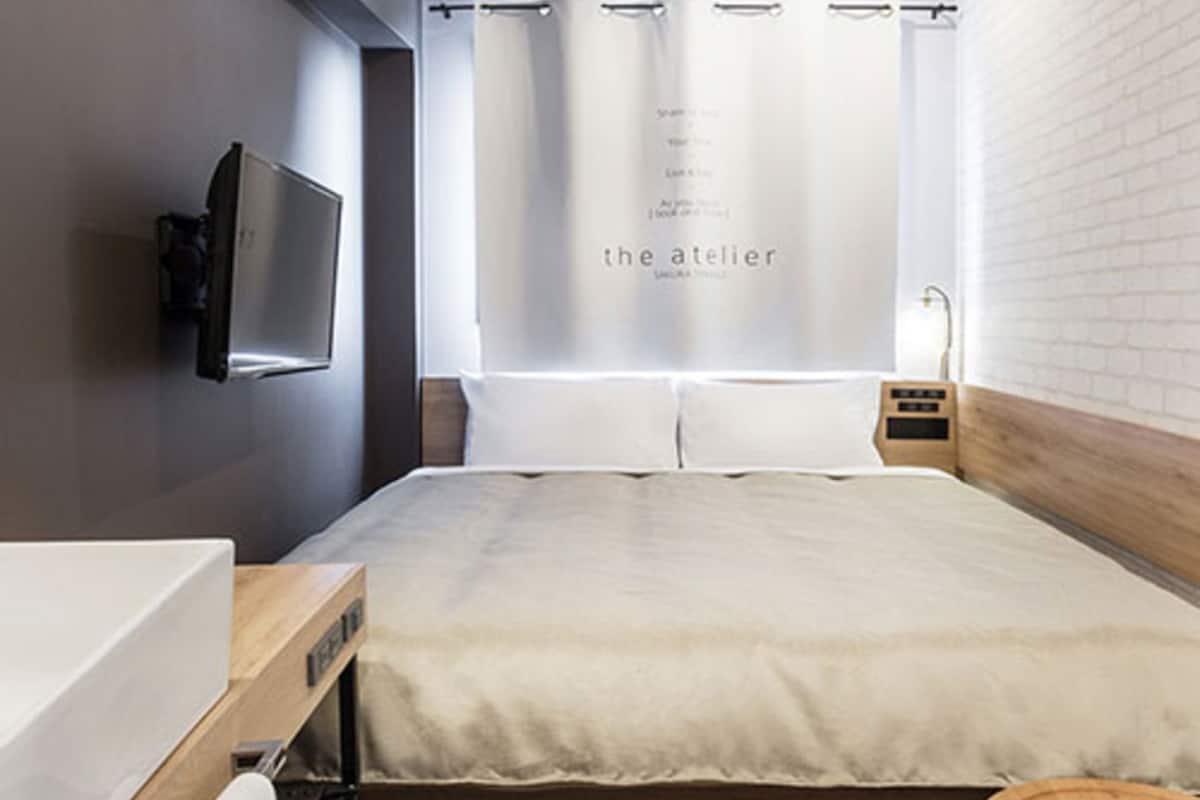 A modern hotel room with a large bed, a TV mounted on the wall, a bedside table with a lamp, and a small wooden stool.