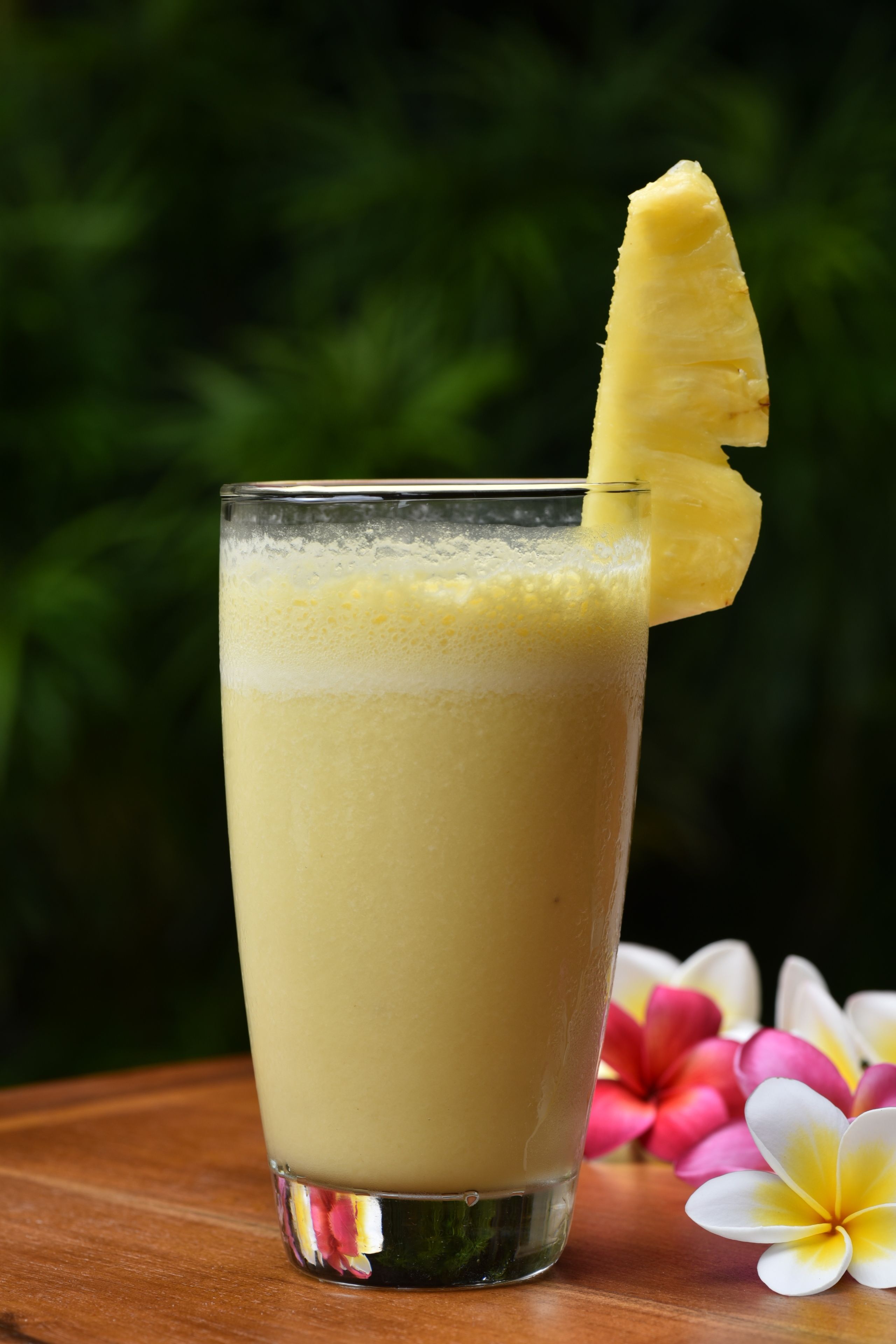 A tall glass with a frothy beverage and a pineapple slice on the rim, placed on a wooden surface with a blurred green background.