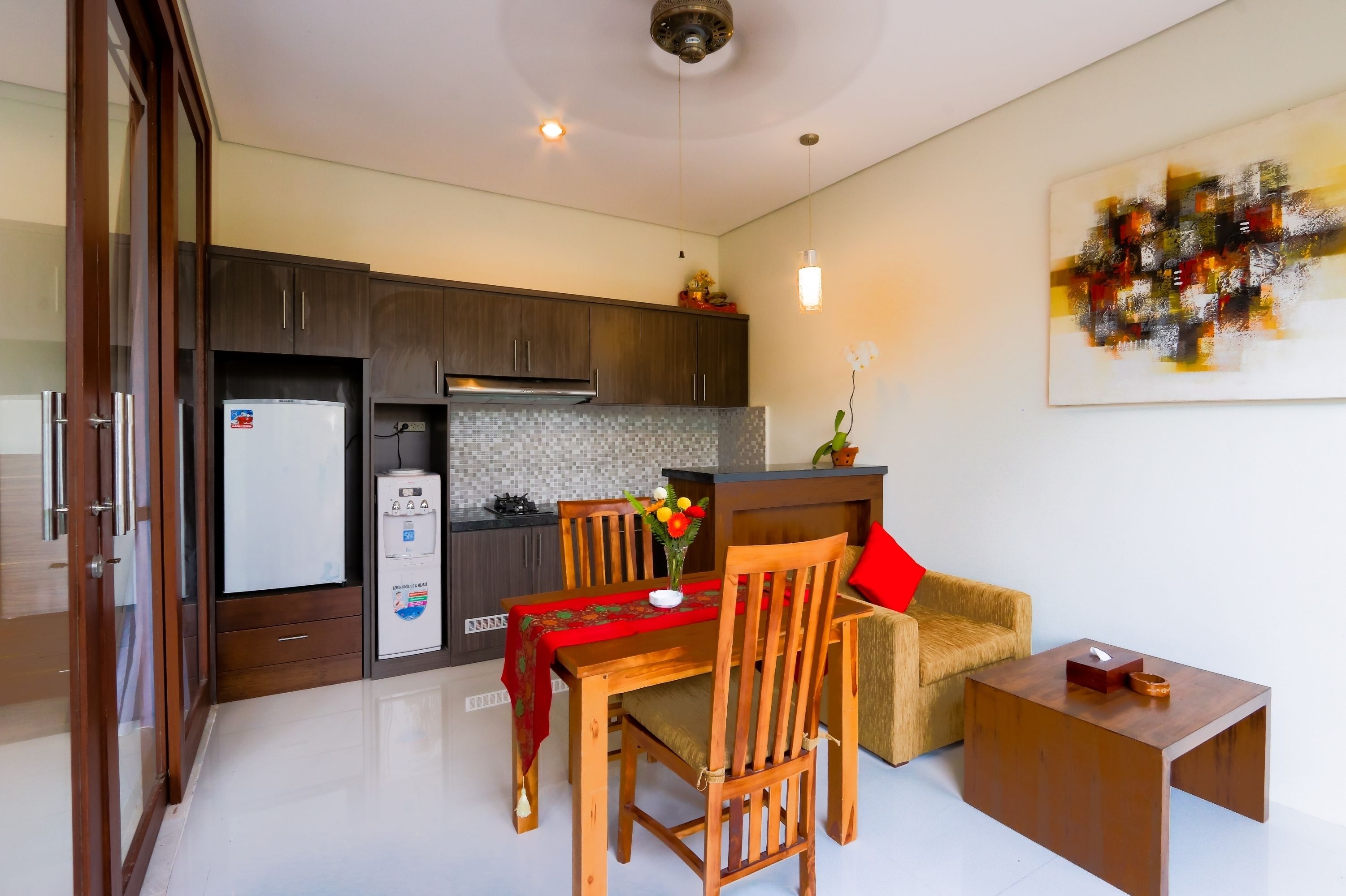 A compact kitchen with dark wood cabinets, a white refrigerator, and a tiled backsplash. A dining table with chairs and a small coffee table is positioned in front of the kitchen.