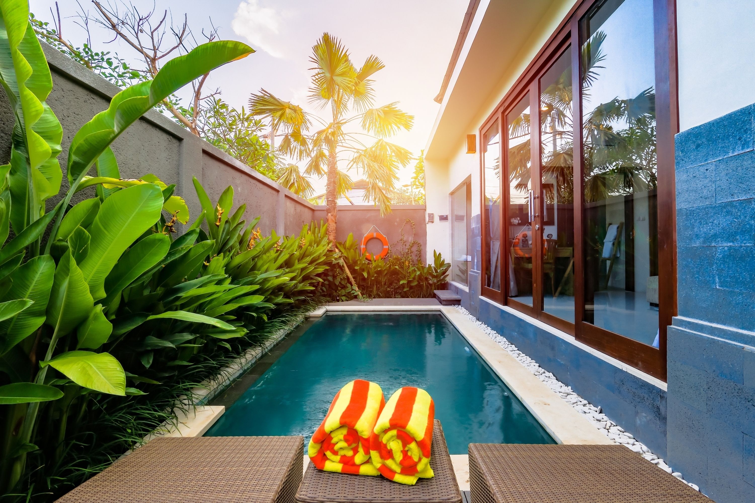 Una piscina con una tumbona y toallas, rodeada de exuberante vegetación y un edificio moderno.