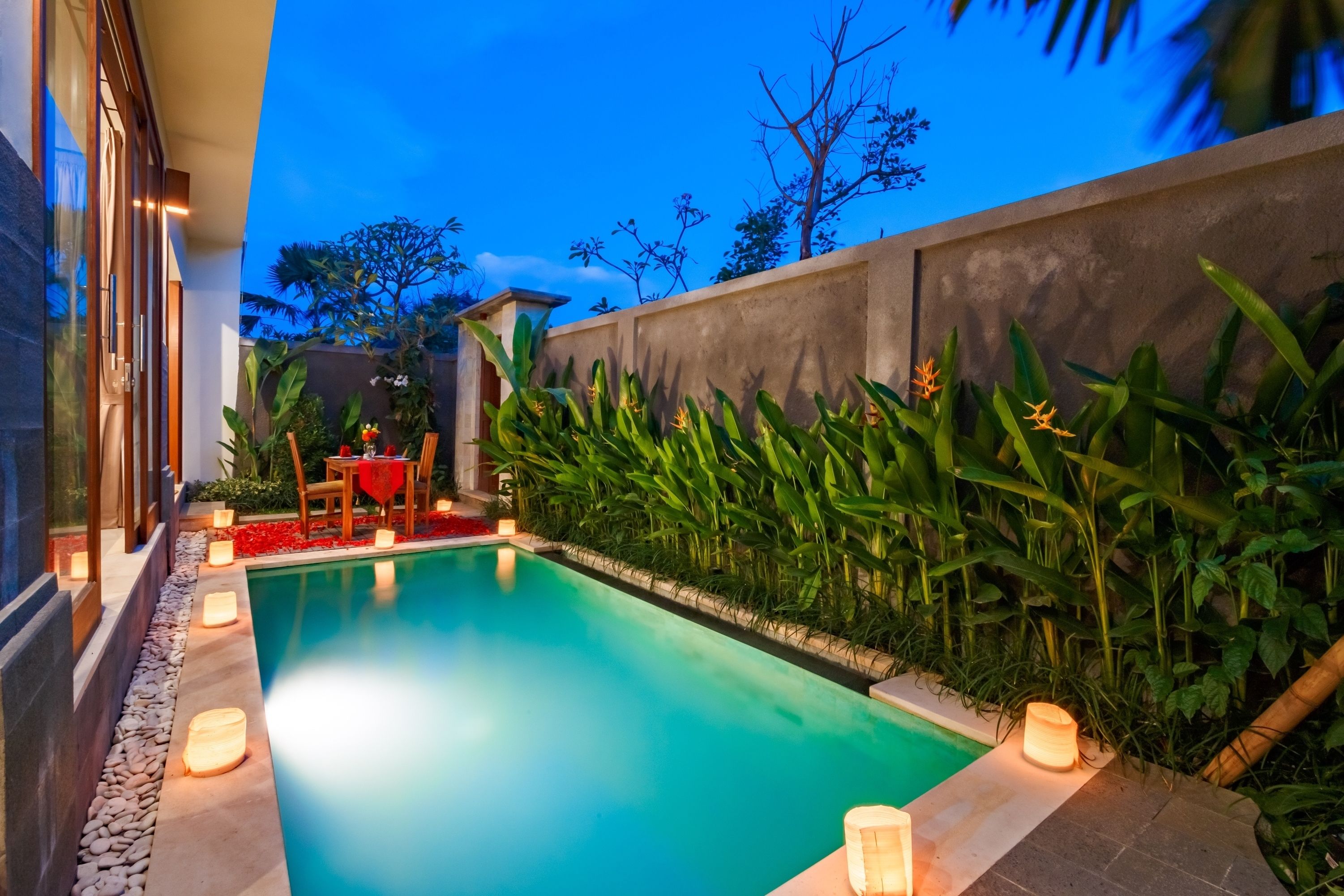 A swimming pool with a stone border, surrounded by lush greenery and a wooden deck with a table and chairs.