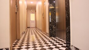 Corredor de hotel com piso xadrez, porta com a inscrição "TOILET" e elevador com paredes de mármore.