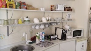Cocina compacta con armarios blancos, un lavamanos de acero inoxidable, microondas y cafetera. Hay repisas sobre la encimera con varios objetos como cajas y frascos.
