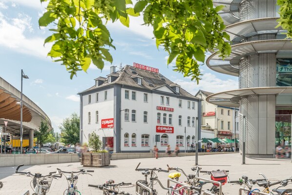A city hotel with a modern design, located in a busy urban area with visible shops and a bicycle parking area. - City Hotel Wetzlar (Wetzlar)