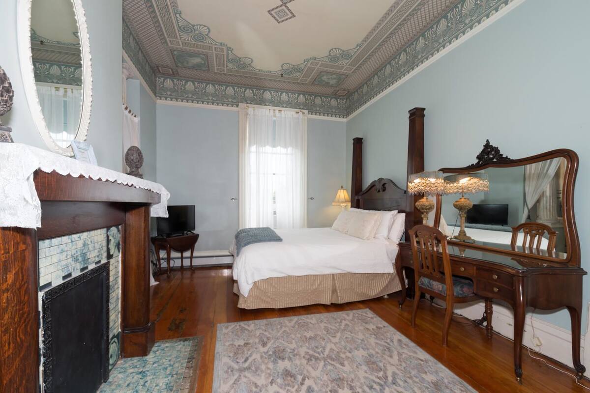 A bedroom with a bed, a desk, a chair, a dresser, a mirror, and a fireplace.