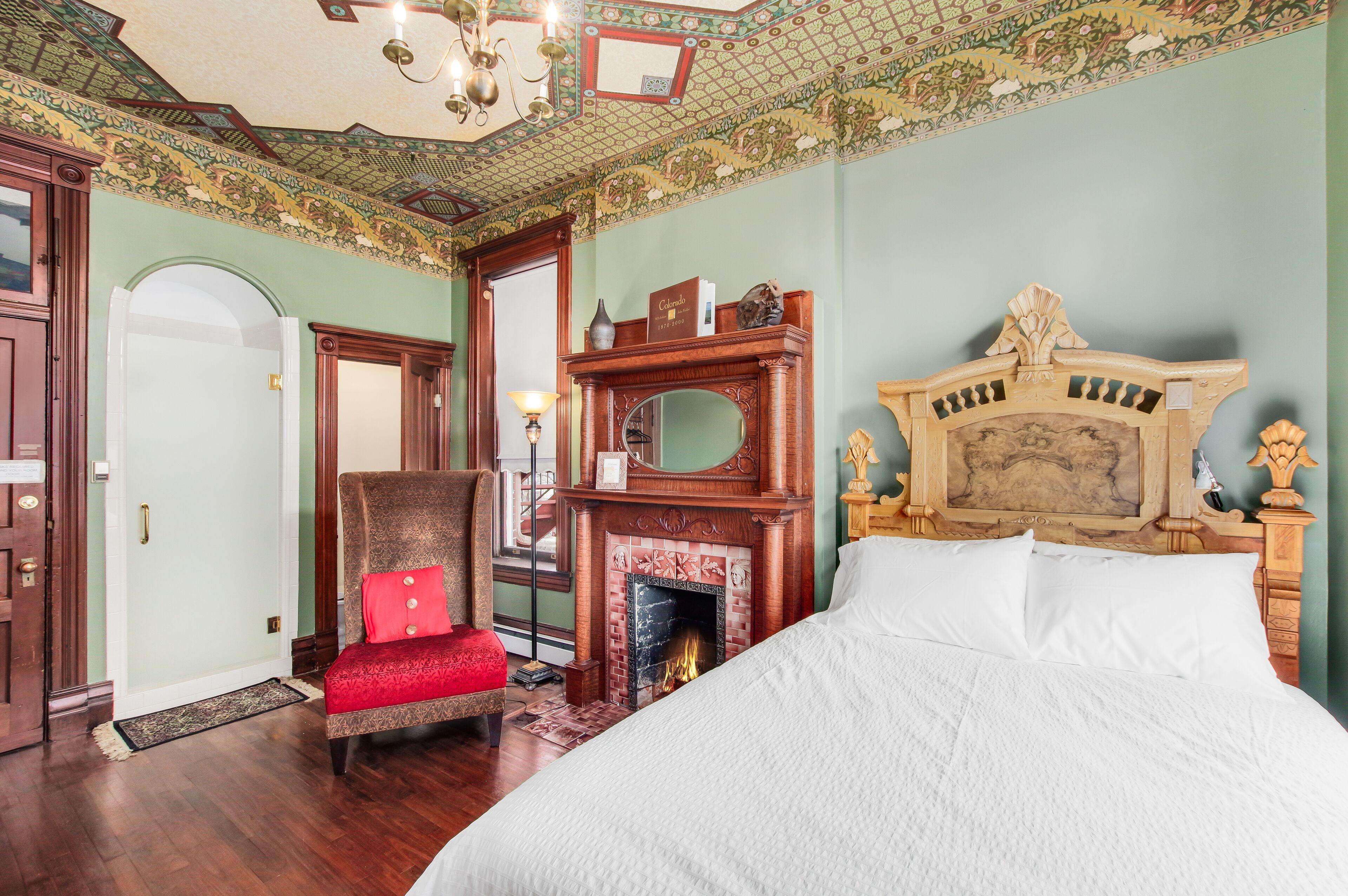 Un dormitorio con una cama grande, una chimenea, un sillón con un cojín rojo y un cabecero de madera.