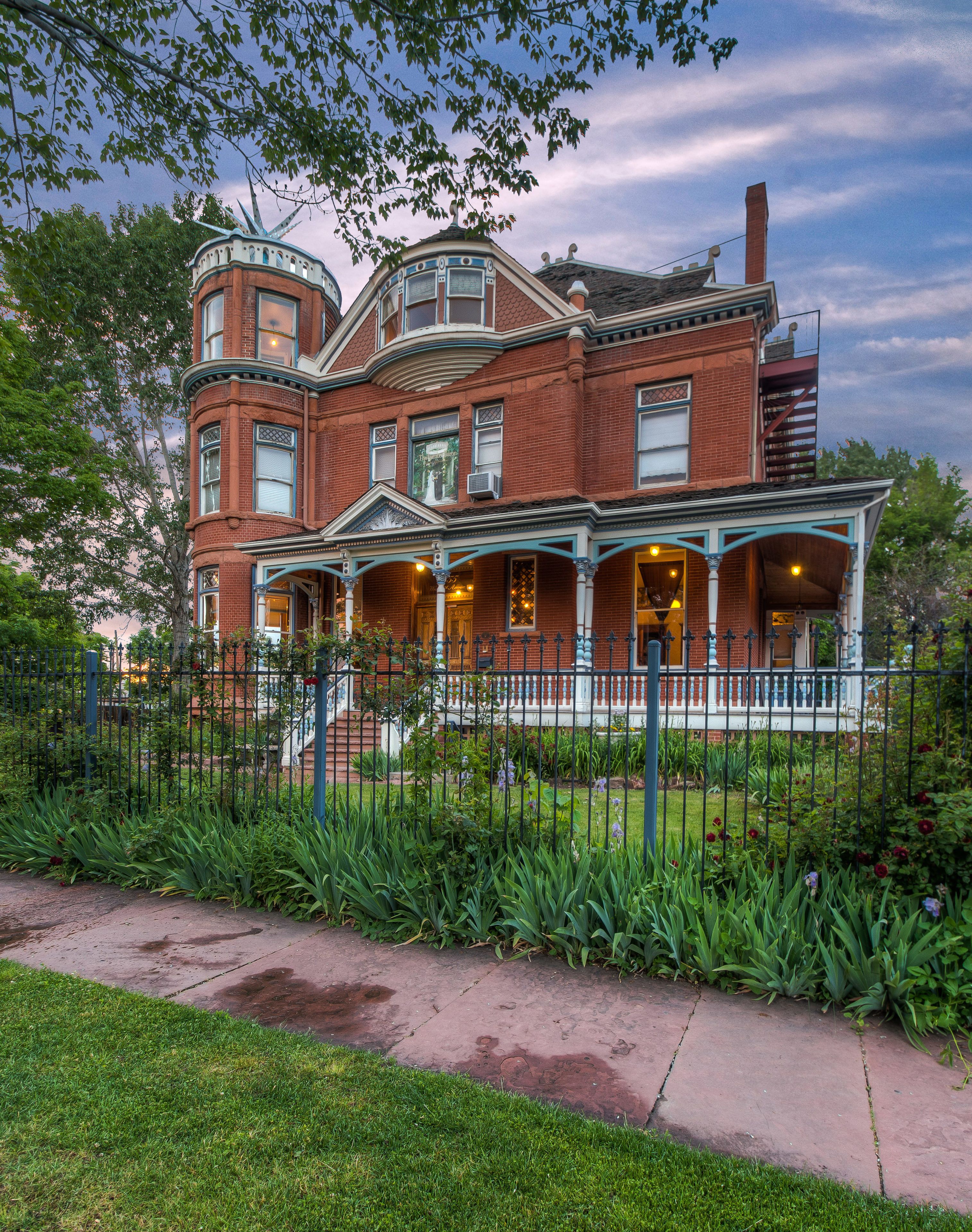 A Victorian-style brick house with a wrap-around porch, a turret, and a large front yard with a fence.