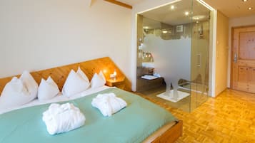 A hotel room with a wooden headboard, a bed with white pillows and towels, a glass-enclosed shower area, and a wooden floor.