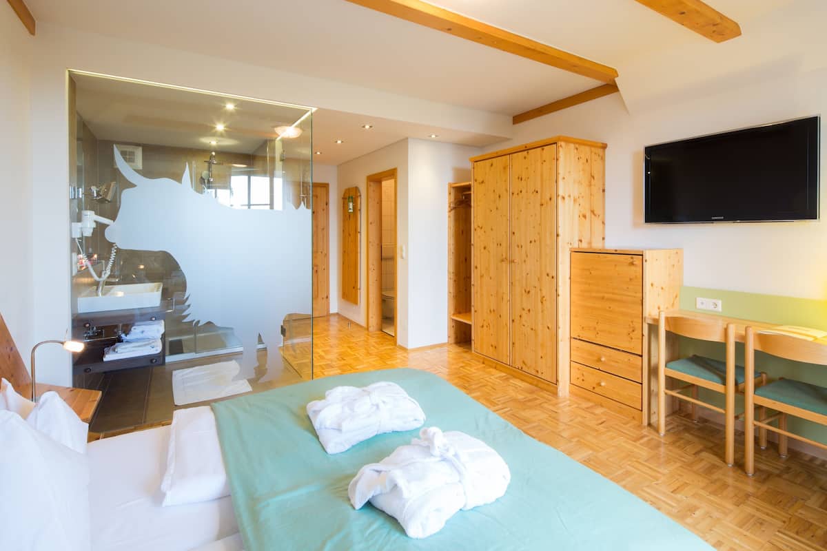Ein Hotelzimmer mit einem Bett, einem Holzschrank, einem Fernseher und einem durch eine Glasscheibe einsehbaren Badezimmer.