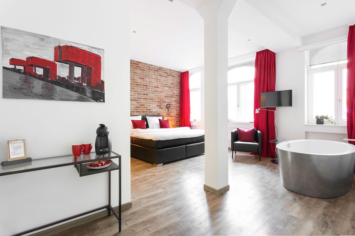 Ein modernes Hotelzimmer mit Backsteinwand, einem Bett mit roten Kissen, einem Couchtisch, einem Fernseher und einer großen Badewanne.