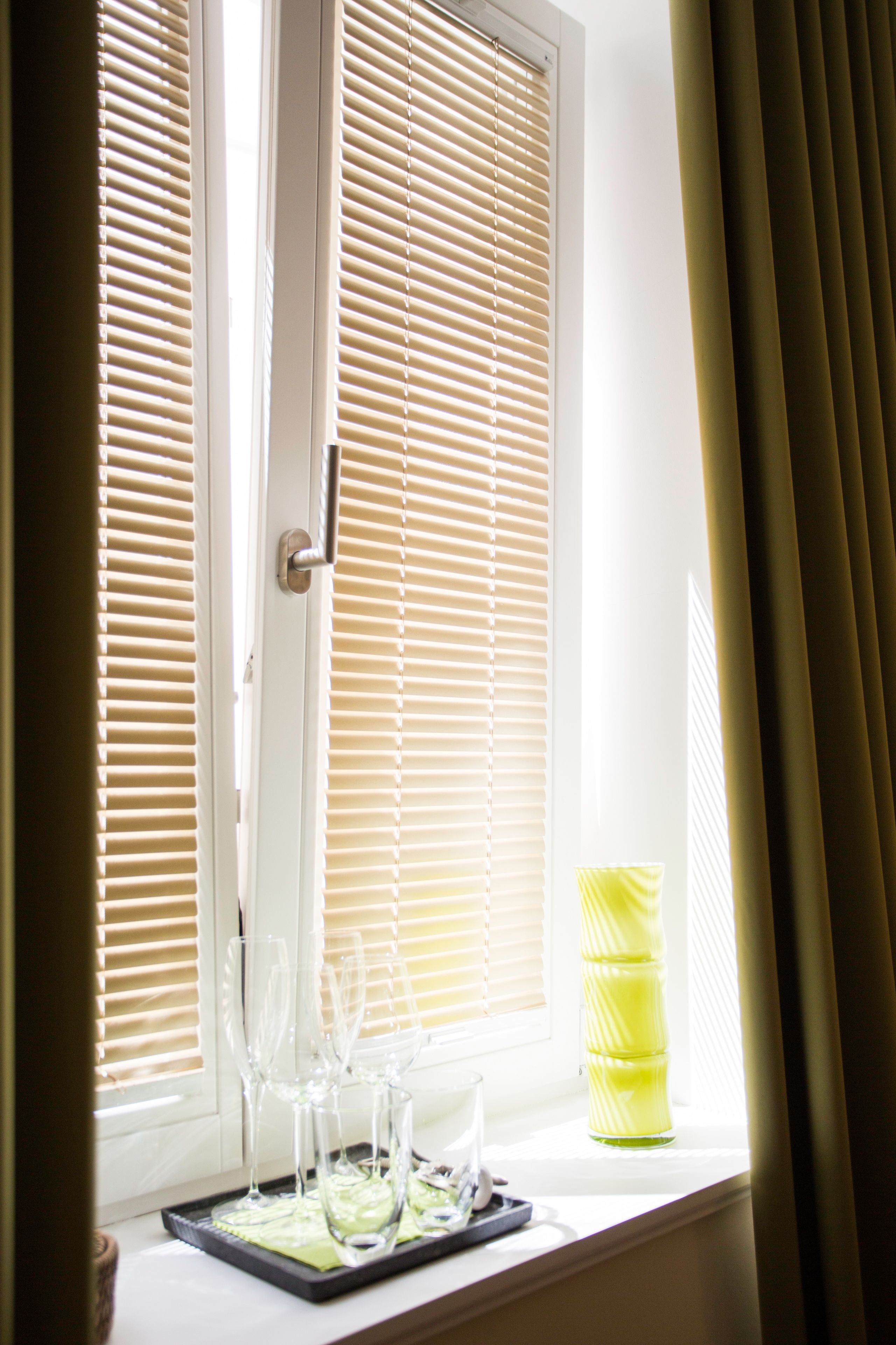 Ein Fenster mit Jalousien, ein Tablett mit Gläsern und eine gelbe Vase.