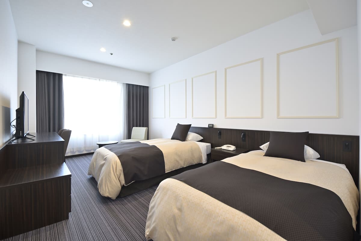 A hotel room with two beds, a desk, a chair, a TV, and a large window with curtains.
