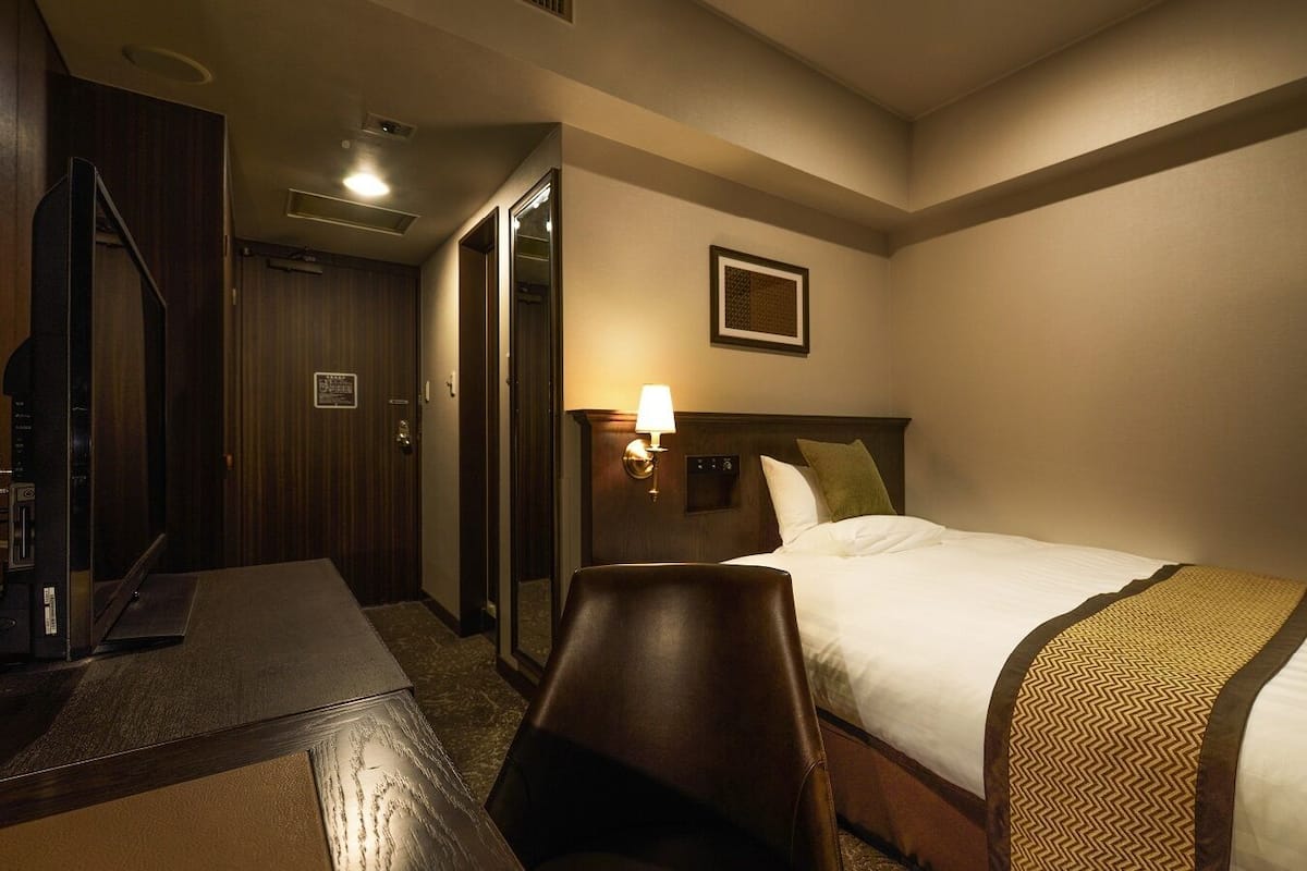 A hotel room with a bed, a desk, a chair, a lamp, and a mirror.