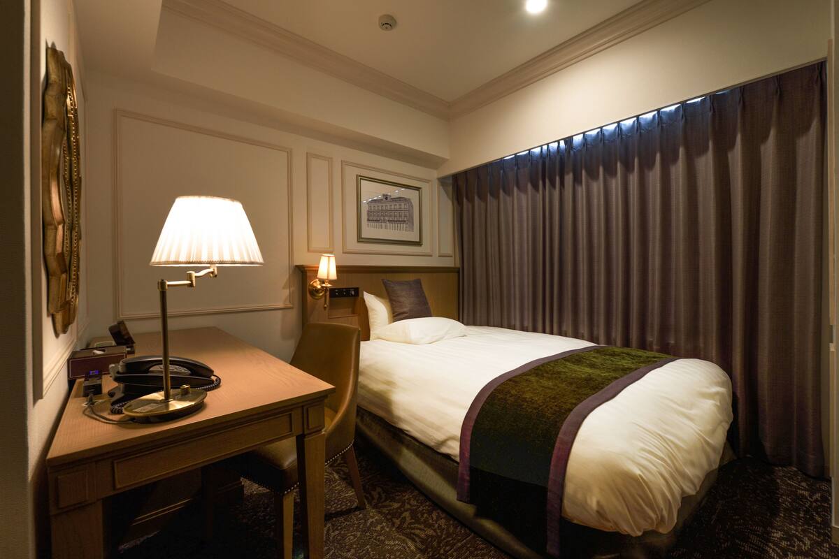 A hotel room with a bed, desk, lamp, and a framed picture on the wall.