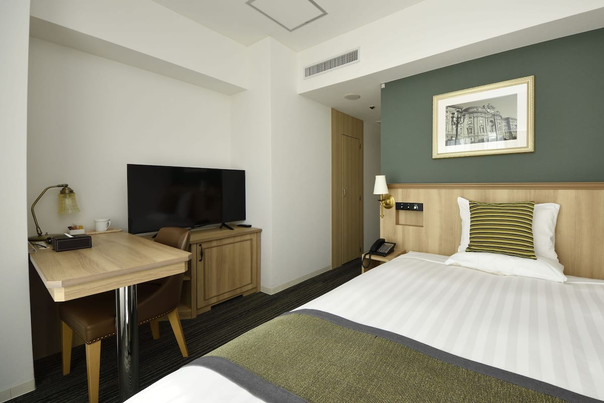 A hotel room with a bed, a desk with a TV, a telephone, and a framed picture on the wall.