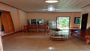 A spacious room with a tiled floor, a wooden bench, a glass-top table with a vase, a refrigerator, and a window with a view of greenery. - Happy Resort (Khanom)