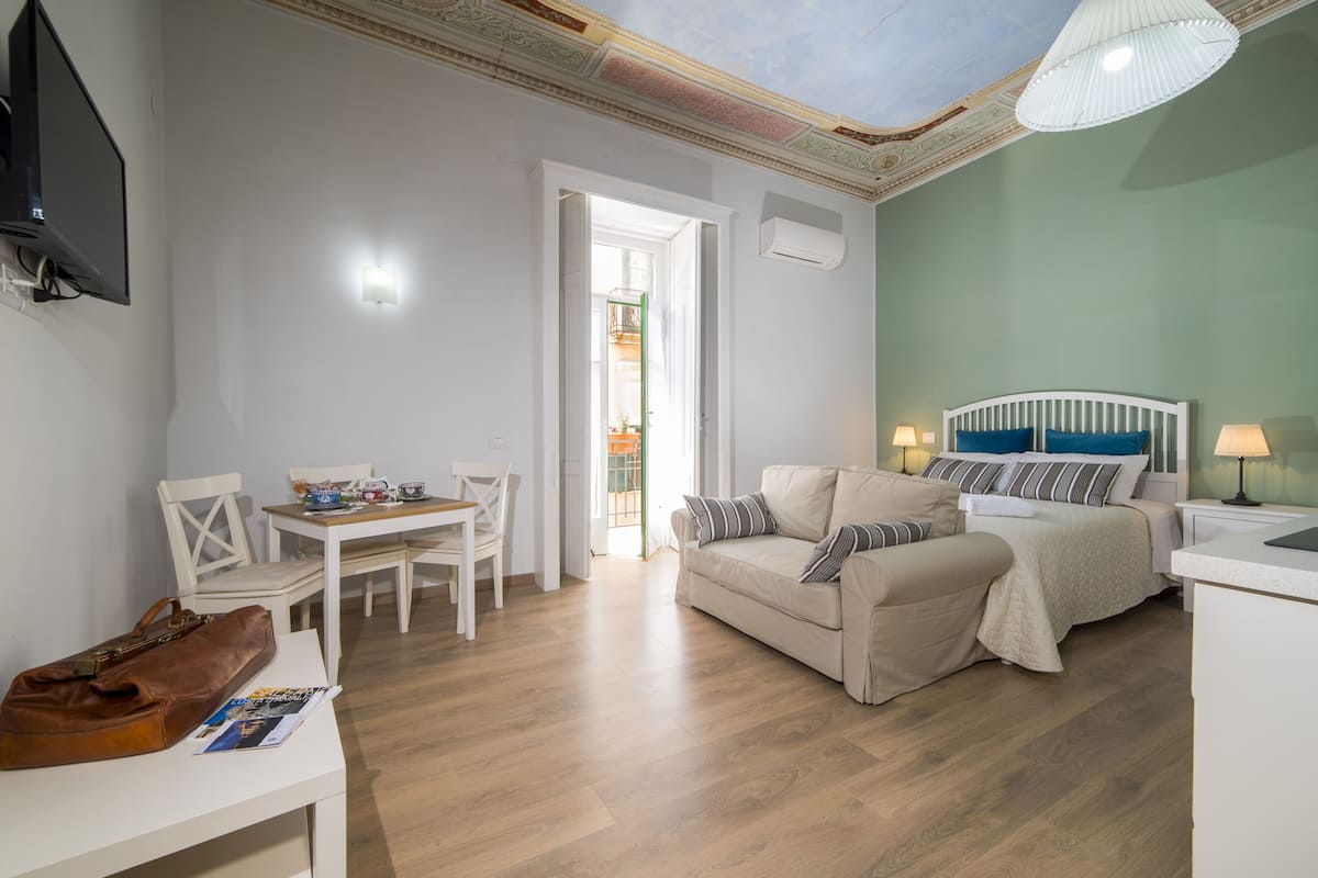 Quarto compacto com cama, sofá, mesa de jantar com cadeiras e TV fixada na parede.