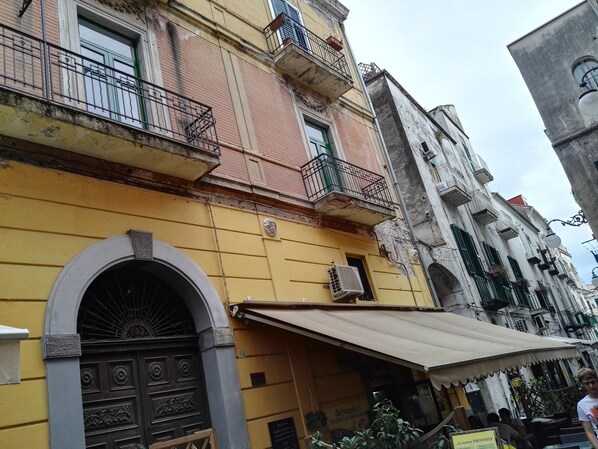 Un edificio amarillo con una gran entrada arqueada, un balcón con barandillas metálicas y un pequeño toldo sobre la entrada.
